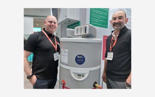 two smiling men on either side of a hot water heat pump